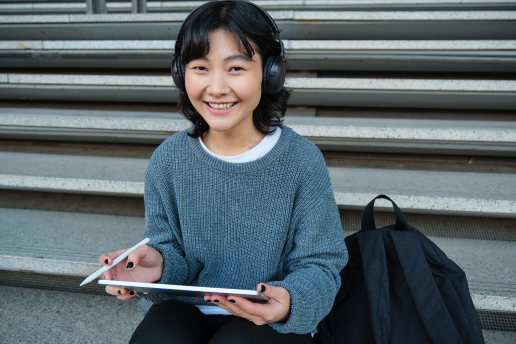 young asian girl, student in headphones, works on remote, digital artist drawing on tablet with graphic pen, listening music in headphones and sitting on street staircase